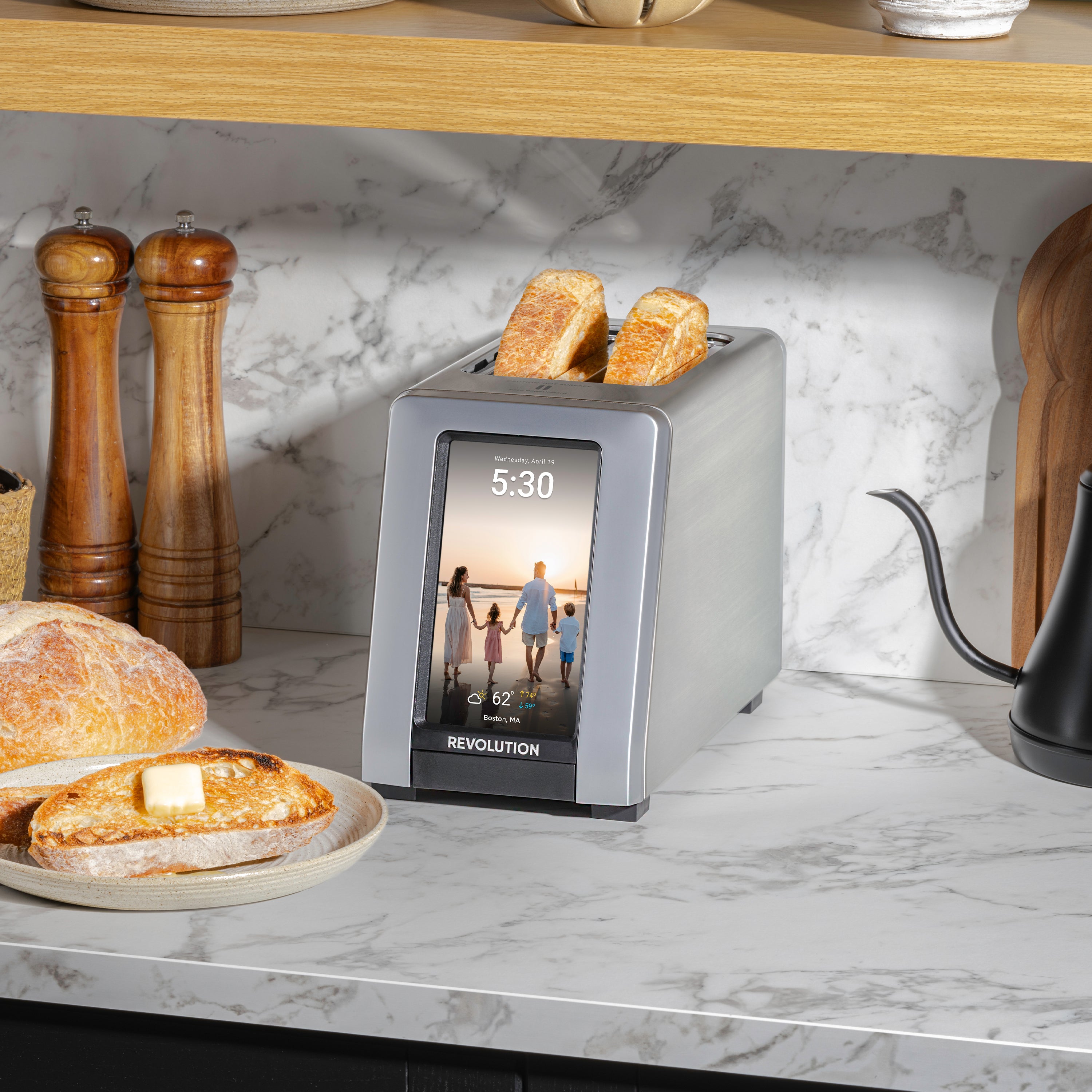 A sleek Revolution Cooking R180 Connect Plus Smart Toaster, featuring a digital screen that shows the time and a family photo, is toasting two slices of bread. It rests on a marble countertop surrounded by buttered toast, a loaf of bread, pepper mills, and a black kettle.