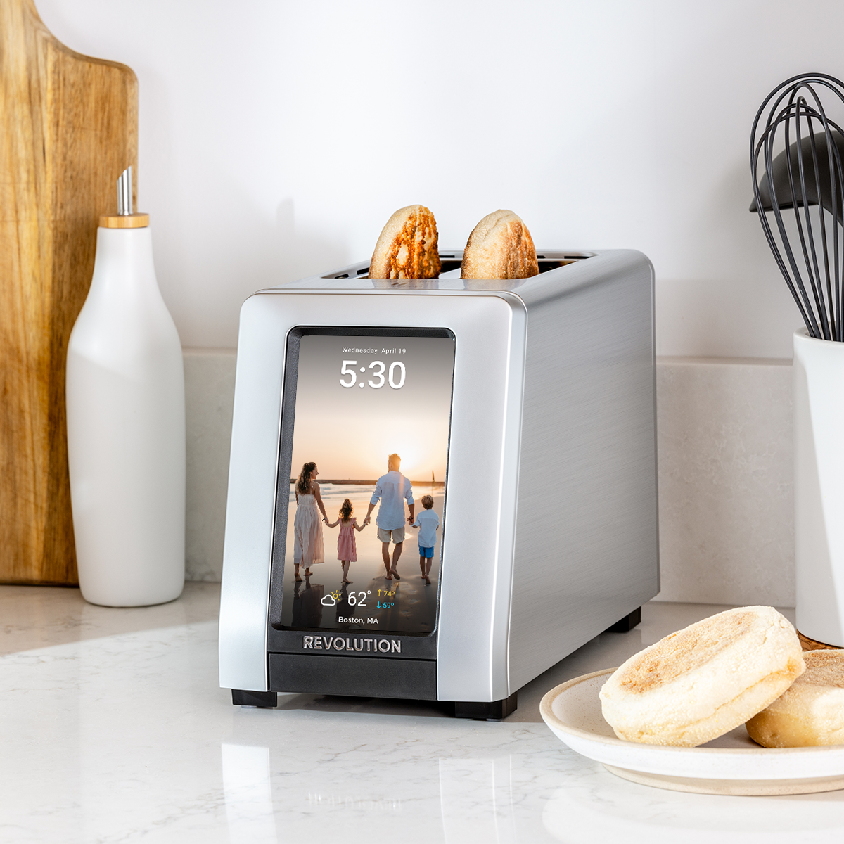 A sleek, modern Revolution Cooking R180 Connect Plus Smart Toaster is stationed on the counter, its touchscreen showing the time, date, weather, and a family photo. Two English muffins are being toasted with precision. Nearby sit a white bottle, cutting board, and kitchen utensils—a seamless mix of technology and style.