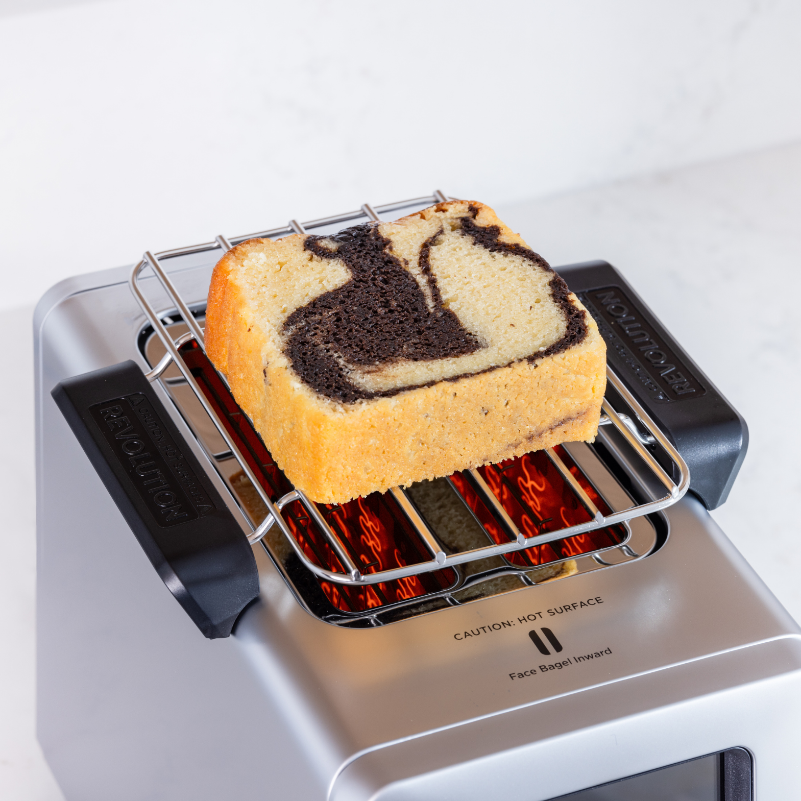 A slice of marble pound cake rests on a stainless steel Warming Rack for Revolution InstaGLO Toasters, with swirls of dark chocolate and light vanilla visible. The warming rack, set against a light background, gently warms the cake while displaying caution labels for hot surfaces.